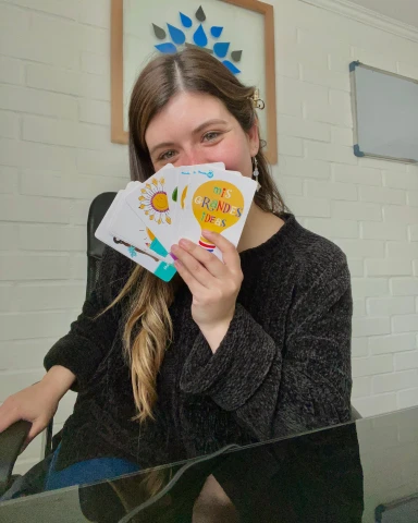 Psícologa Paz sonriendo, sentada en una silla de oficina y sosteniendo un abanico de tarjetas coloridas con ilustraciones y texto, presumiblemente para uso terapéutico.