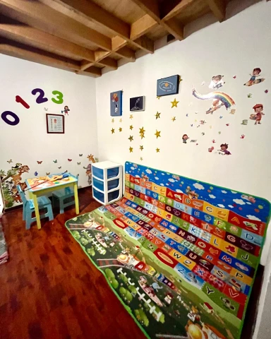 Una habitación diseñada para terapia infantil con un piso de madera oscura, paredes blancas decoradas con números, estrellas y dibujos animados. Hay una mesa pequeña con dos sillas de colores, un mueble de almacenamiento y una gran alfombra de juego con el abecedario.